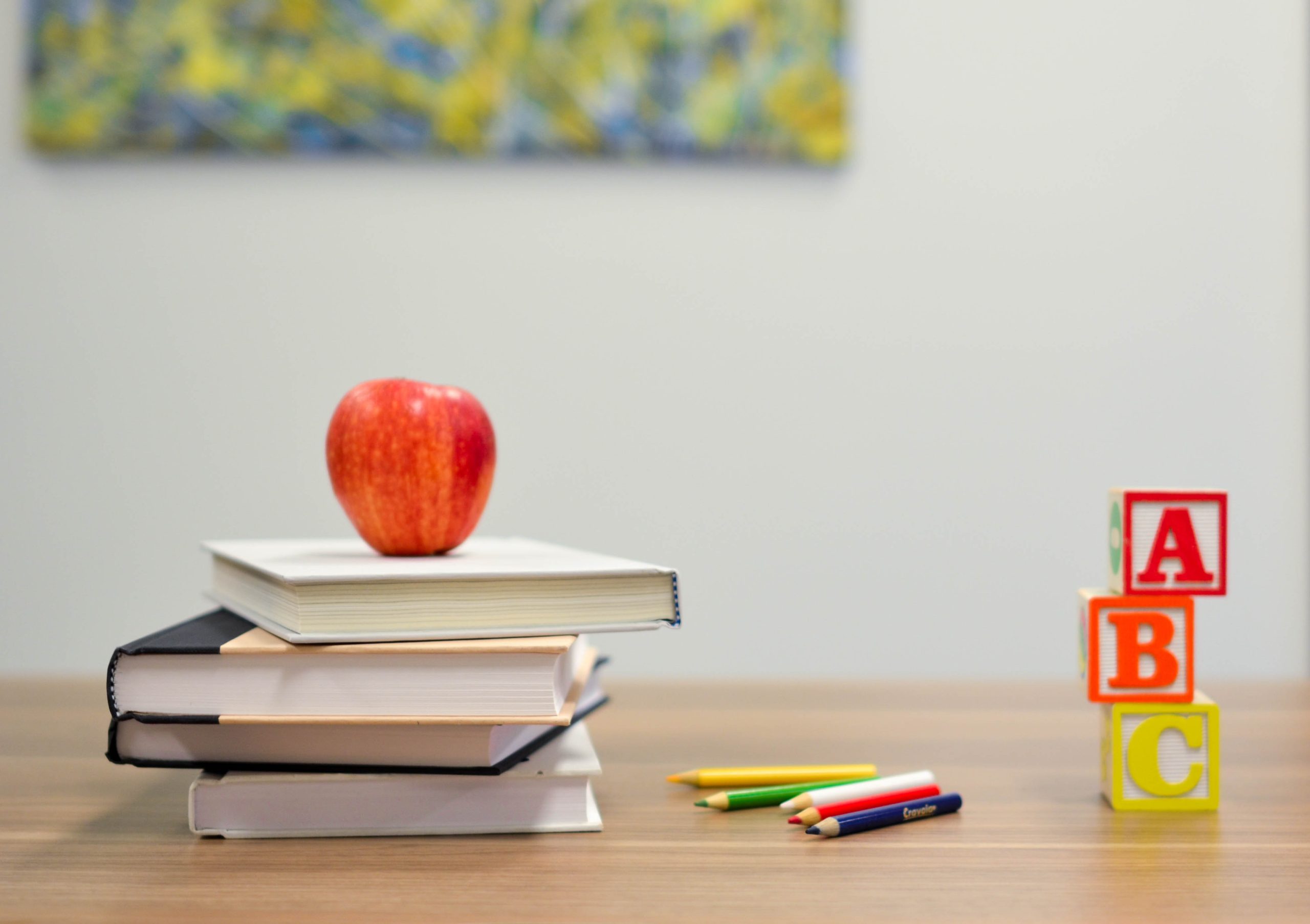 Pile of books with an apple on top in a classroom. Pencils and ABC blocks are visible